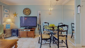A dining area with a round table, chairs, a television, and a clock on the wall. - Makin' Memories @ Victorian (GVR05004) (Galveston)