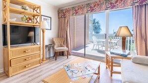 A living room with a TV, a sofa, a coffee table with board games, a chair, and a balcony with a table and chairs.