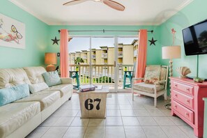 Quarto com sofá bege, cómoda rosa, televisão e varanda com vista para edifícios.