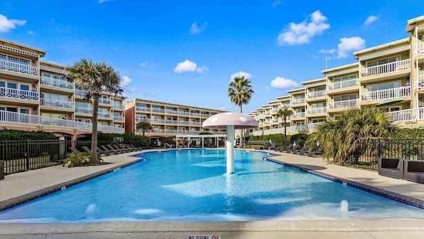 Piscina com um aviso de 'NÃO MERGULHAR', rodeada por um hotel com varandas e palmeiras.