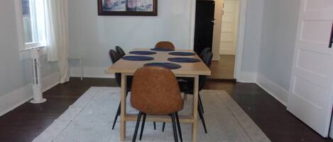 Een eethoek met een tafel en stoelen, een wit vloerkleed, een schilderij aan de muur en een deur op de achtergrond.
