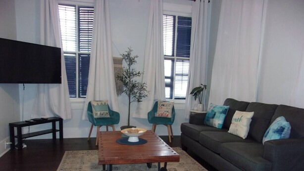 A living room with a dark wood coffee table, two teal chairs, a gray sofa with blue and white cushions, and a large window with white curtains.