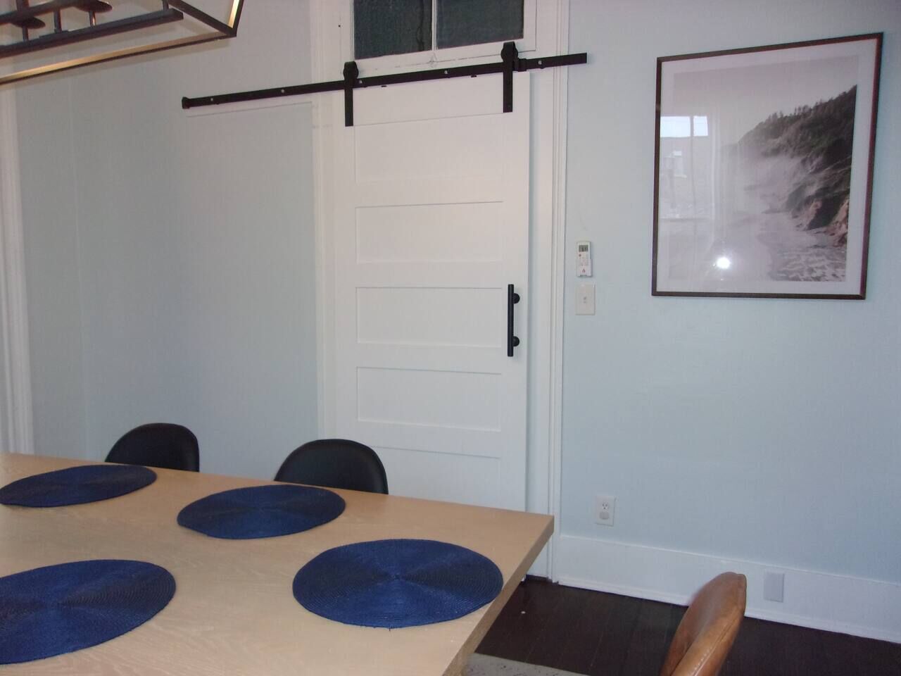 Une pièce avec une table en bois, quatre chaises, une barre horizontale noire, une porte blanche et un tableau encadré au mur.