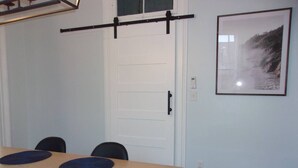 A room with a wooden table, four chairs, a black horizontal bar, a white door, and a framed picture on the wall.