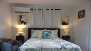 A bedroom with a bed, two bedside tables, a chair, an air conditioning unit, and a wall-mounted picture.