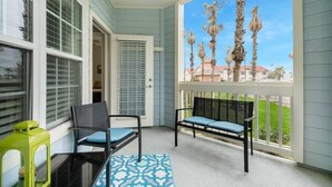 Un balcon avec deux chaises et une table, une porte et une vue sur des palmiers et des maisons.
