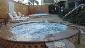 Un jacuzzi con terraza de madera y sillas reclinables, rodeado por una valla de madera y un edificio al fondo.