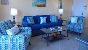 A living room with a blue sofa set, a coffee table, and patterned chairs. There are two wall-mounted lamps and a painting of a van on the wall. - Surf's Up at the Dawn BEACH FRONT (Galveston)