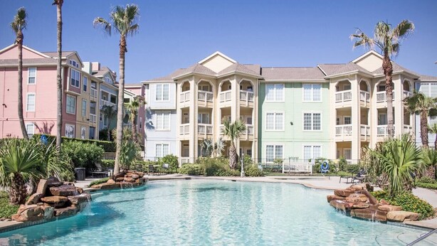 A swimming pool surrounded by multi-story buildings with balconies, palm trees, and a clear sky. - Surf's Up at the Dawn BEACH FRONT (Galveston)