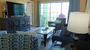 A living room with a television, patterned furniture, a glass coffee table, and a view through a window. - Surf's Up at the Dawn BEACH FRONT (Galveston)