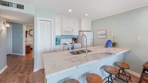 A modern kitchen with white cabinets, a marble countertop, and a sink. There are two stools at the counter and a small island. - Welcome Inn at Maravilla! (Galveston)