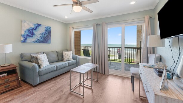 A modern living room with a grey sofa, a small table, and a ceiling fan. There is a large window with curtains, a flat-screen TV, and a painting on the wall. - Welcome Inn at Maravilla! (Galveston)