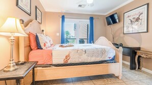 Un dormitorio con cama, mesita de noche, lámpara, ventilador de techo, televisión y vista al balcón.