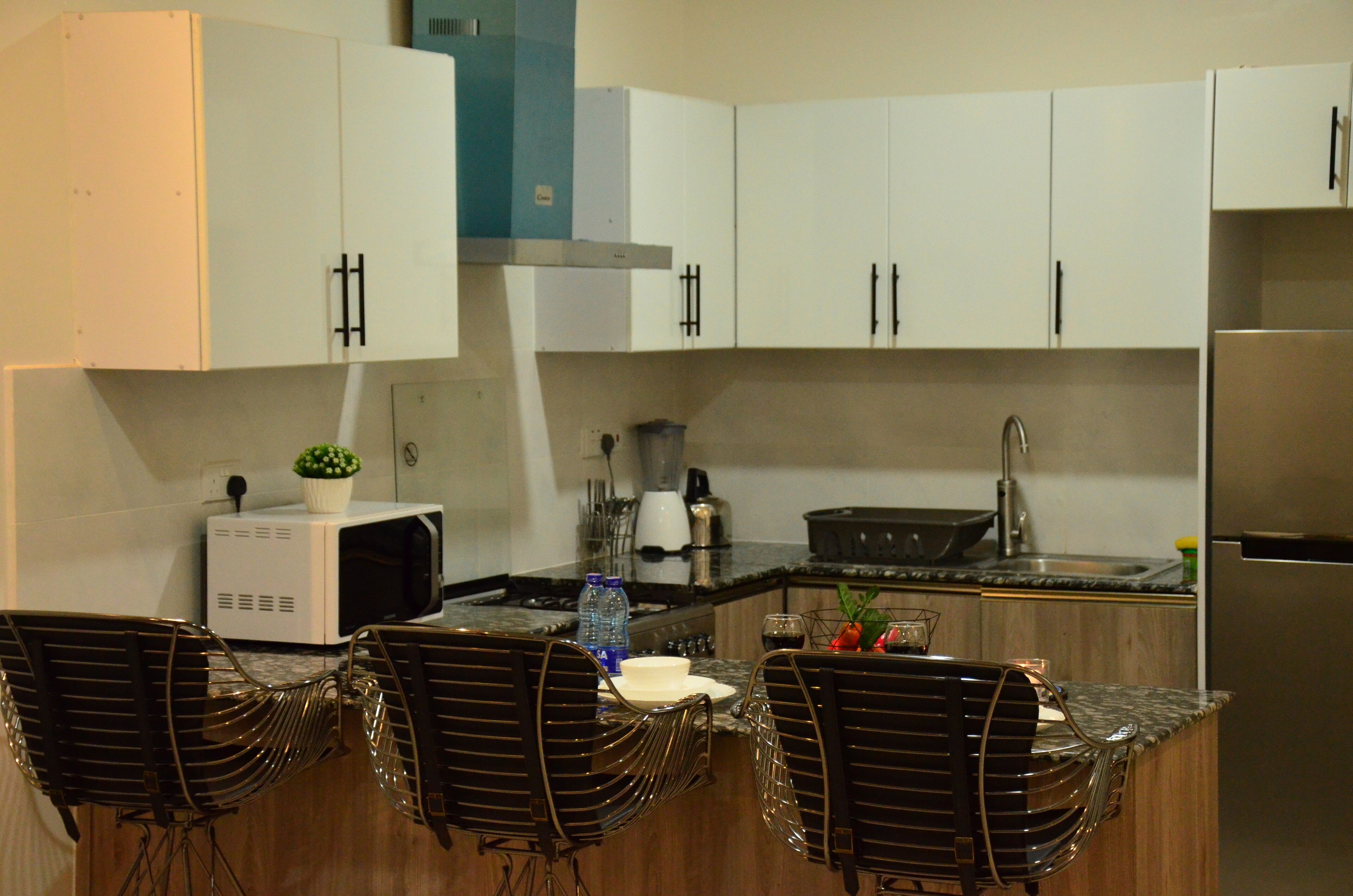 A modern kitchen with white cabinets, a stainless steel refrigerator, and a granite countertop. There are three black chairs with a unique design.