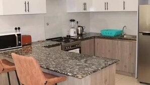 A kitchen with white cabinets, a granite countertop, a stove, a sink, and two bar stools. - Yohana Residences (Nairobi)