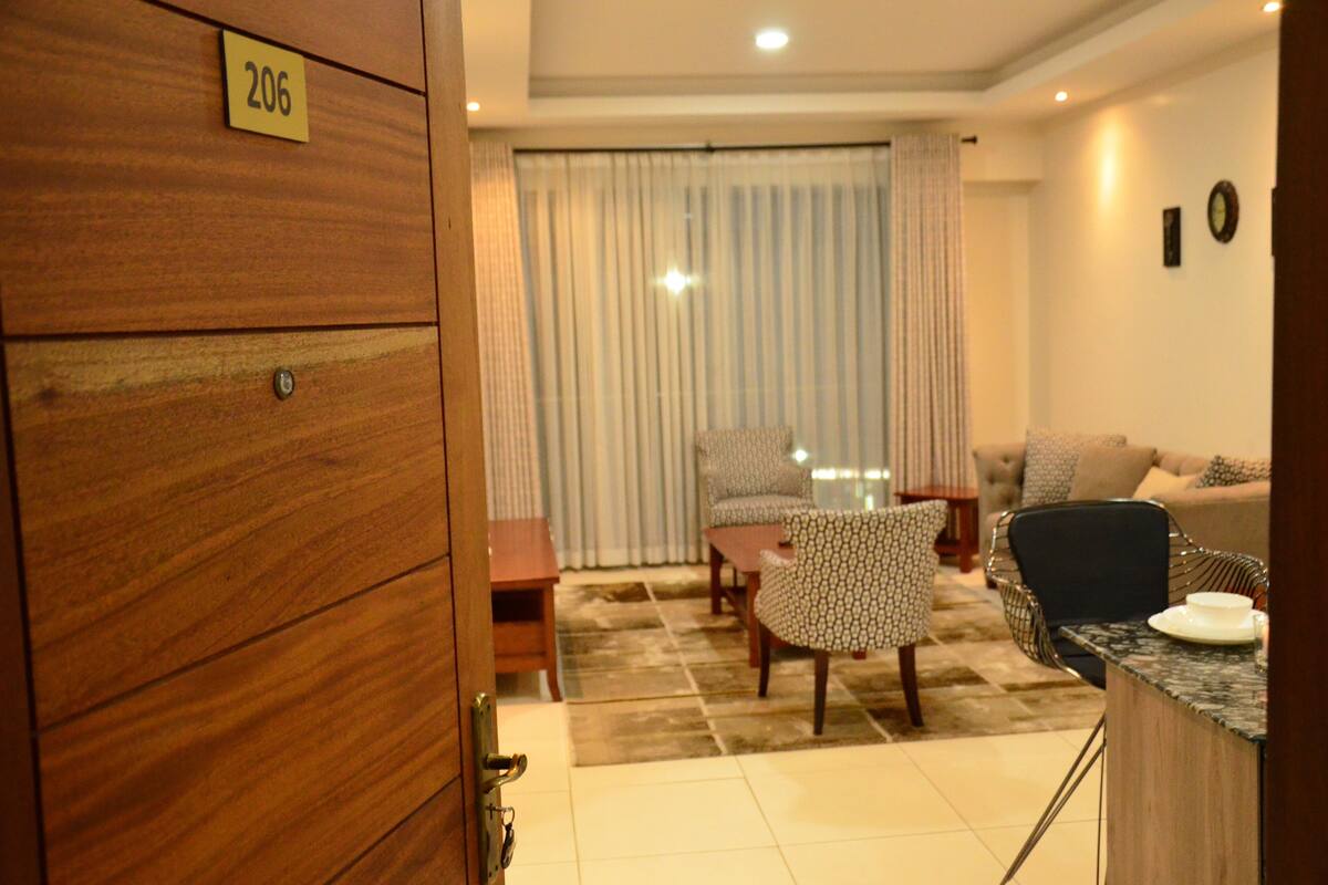 A room with a wooden door labeled '206', a dining table with chairs, a sofa, and a small kitchen area with a marble countertop.