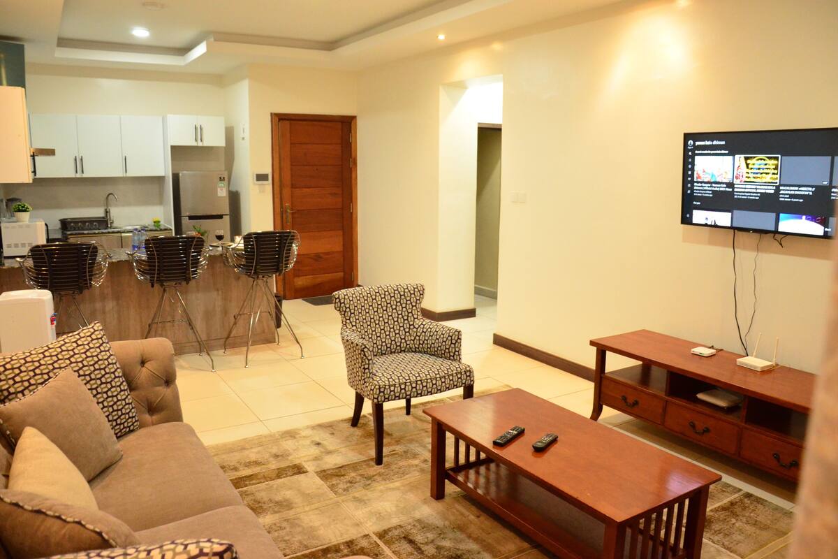 A living room with a sofa, armchair, coffee table, and TV. There is a kitchen in the background.