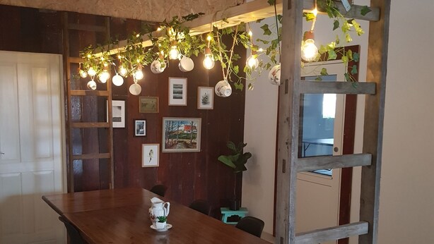 Un comedor con mesa de madera, sillas negras y una escalera. Hay cuadros en la pared y luces colgantes sobre la mesa.