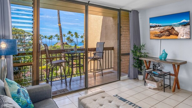 Ein modernes Wohnzimmer mit einer Couch, einem Hocker und einem Essbereich mit Stühlen. Ein Fernseher ist an der Wand montiert und es gibt einen Balkon mit Blick auf Palmen und das Meer.