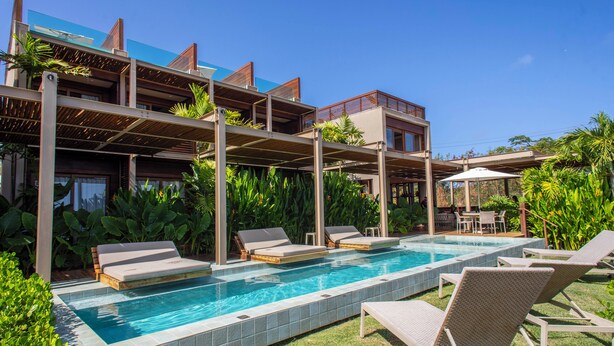 A modern pool area with lounge chairs and a clear blue pool, surrounded by lush greenery and a multi-story building with balconies. - POUSADA DO MIRANTE (Fernando de Noronha)
