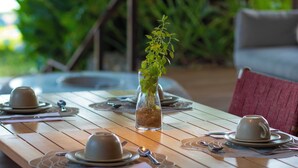 A wooden dining table set with plates, cups, and cutlery, surrounded by chairs, with a potted plant in the center. - POUSADA DO MIRANTE (Fernando de Noronha)