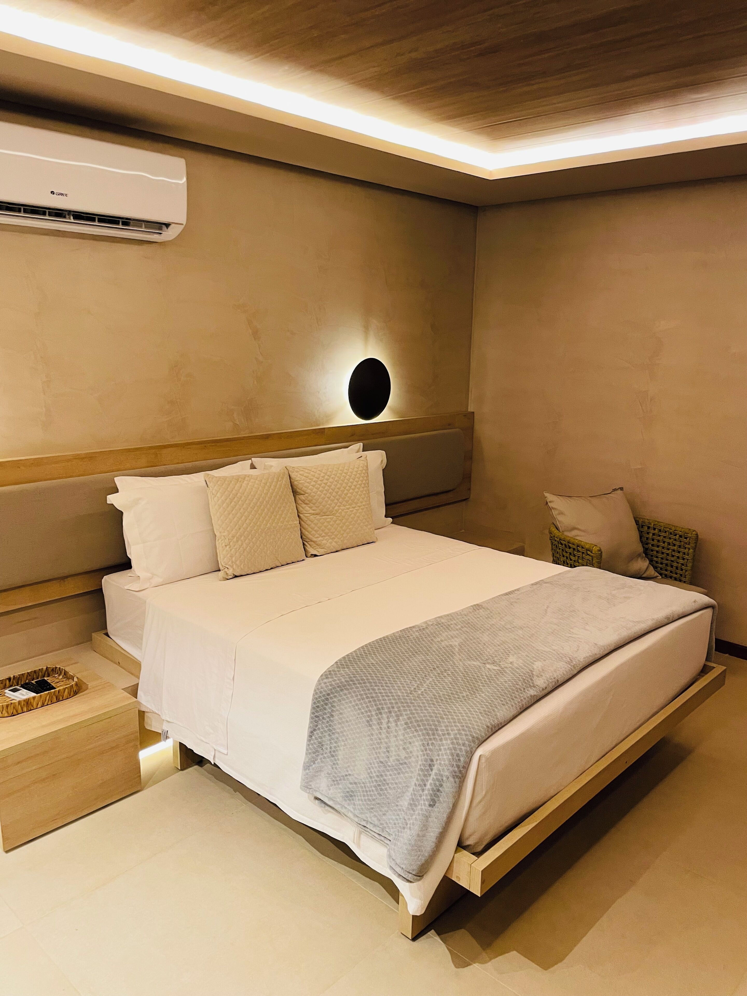 Uma cama bem arrumada com cabeceira, uma mesinha de cabeceira com bandeja e um ar condicionado fixado na parede.