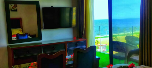 A hotel room with a large TV, a bed, a desk, and a balcony with a view of the sea.