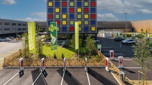 Un edificio moderno con una fachada de ventanas de colores dispuestas en cuadrícula, un estacionamiento con puntos de carga para vehículos eléctricos y lonas publicitarias de un hotel.