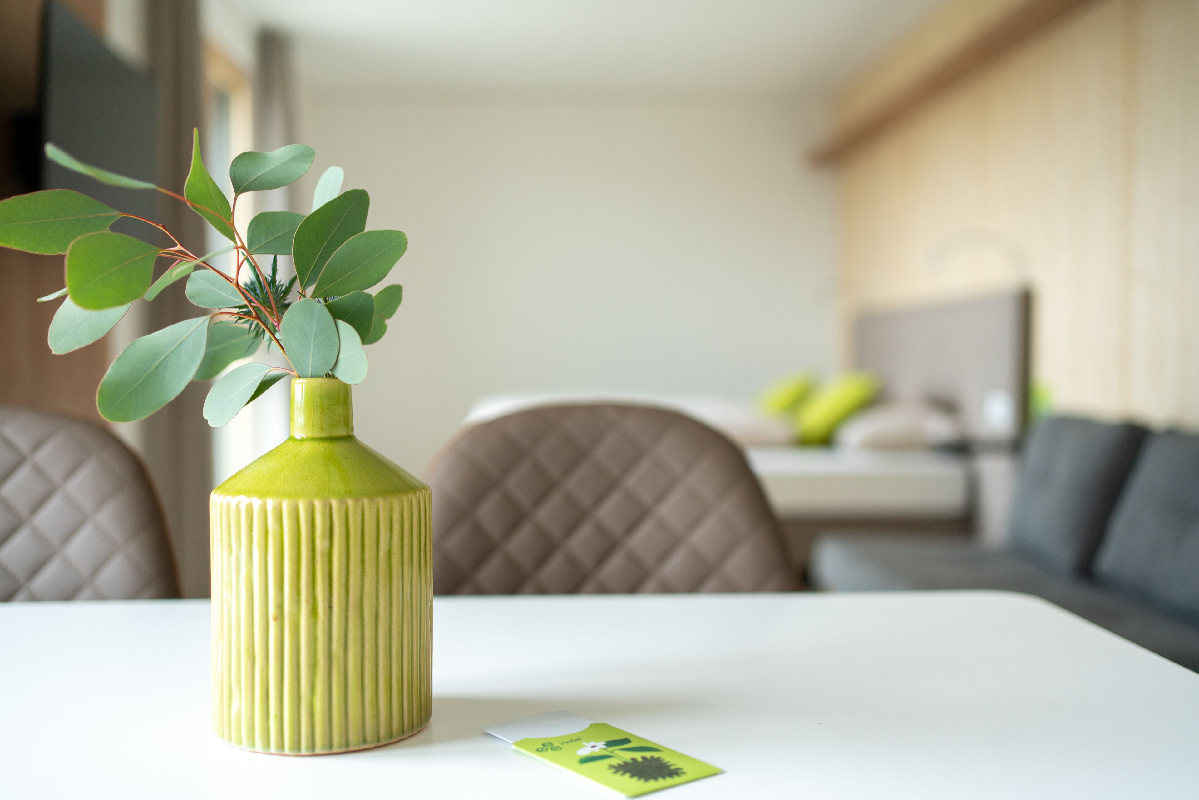 Eine grüne Vase mit Blättern auf einem wei ßen Tisch in einem modernen Raum mit einer Couch und einem Essbereich.