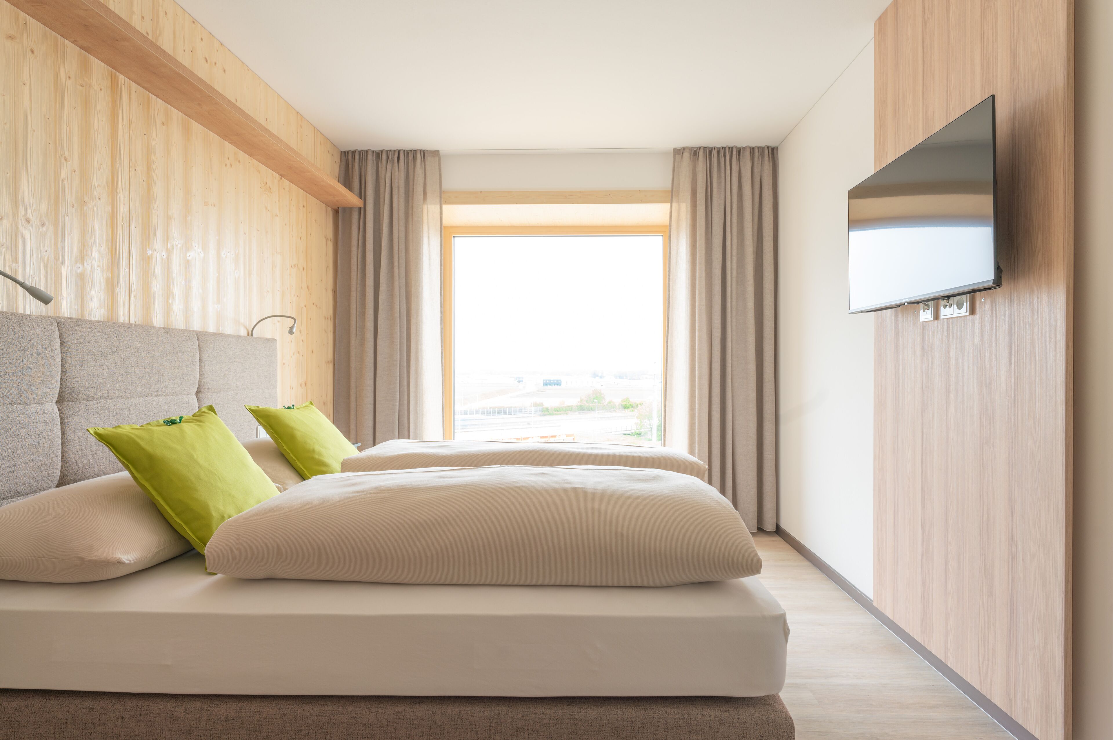 A modern hotel room with a large bed, a flat-screen TV mounted on the wall, and a large window with curtains.