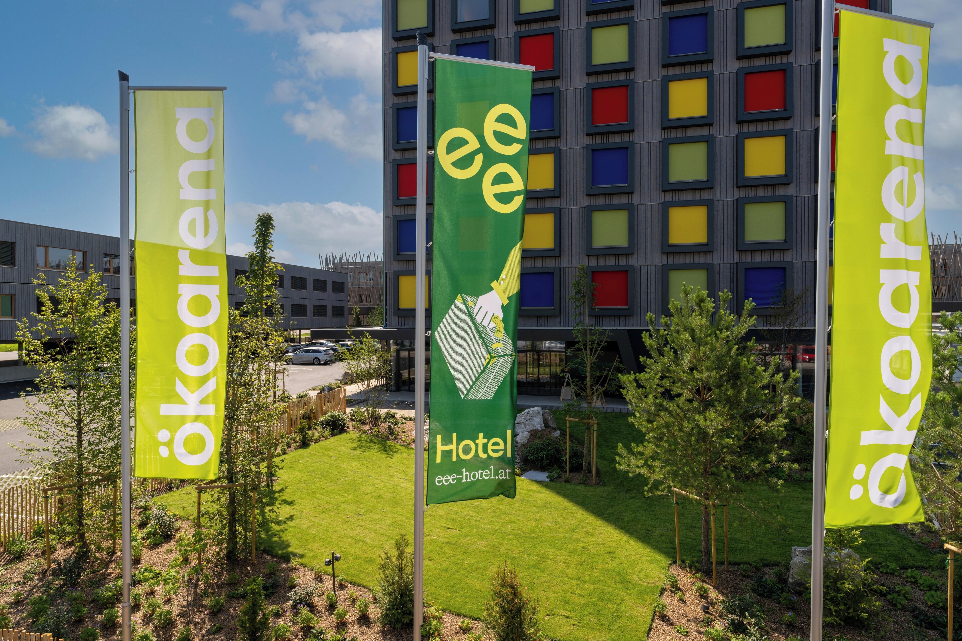 Three flags with the text "ekoarena" and "Hotel eee-hotel.at" in front of a modern building with colorful windows.