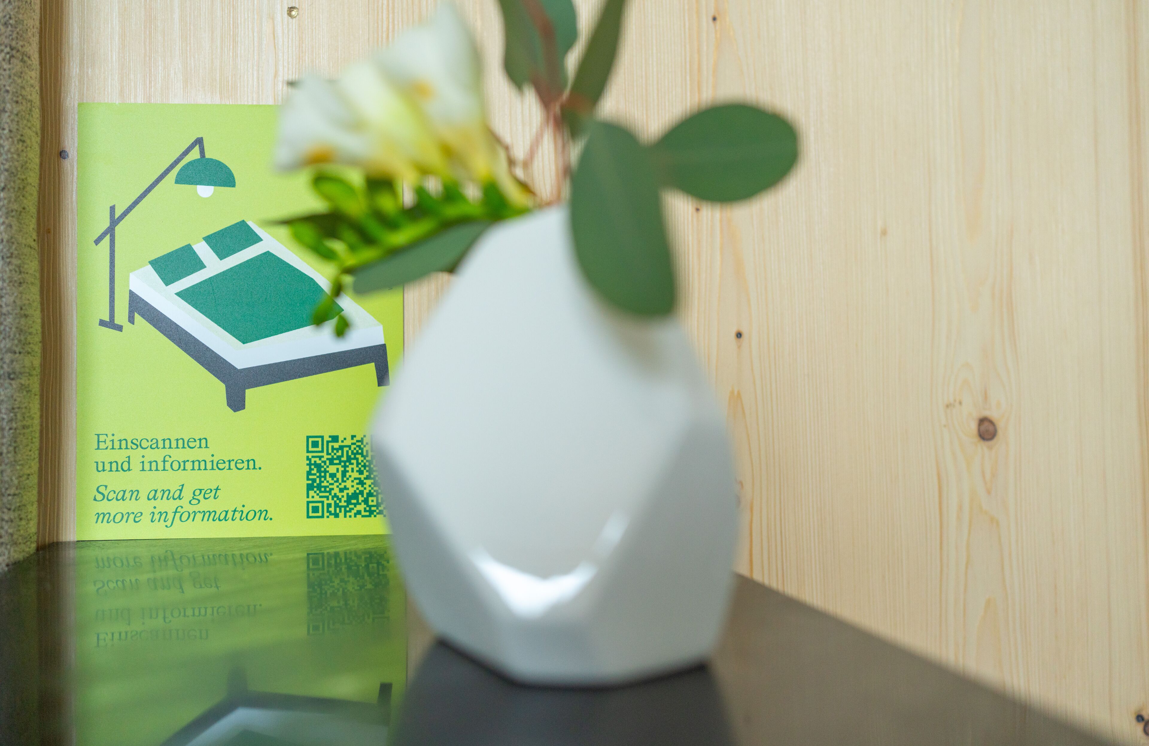 En vit vas med gröna blad står på en träyta, bredvid en QR-kod och en affisch med en illustration av en säng och tillhörande text.