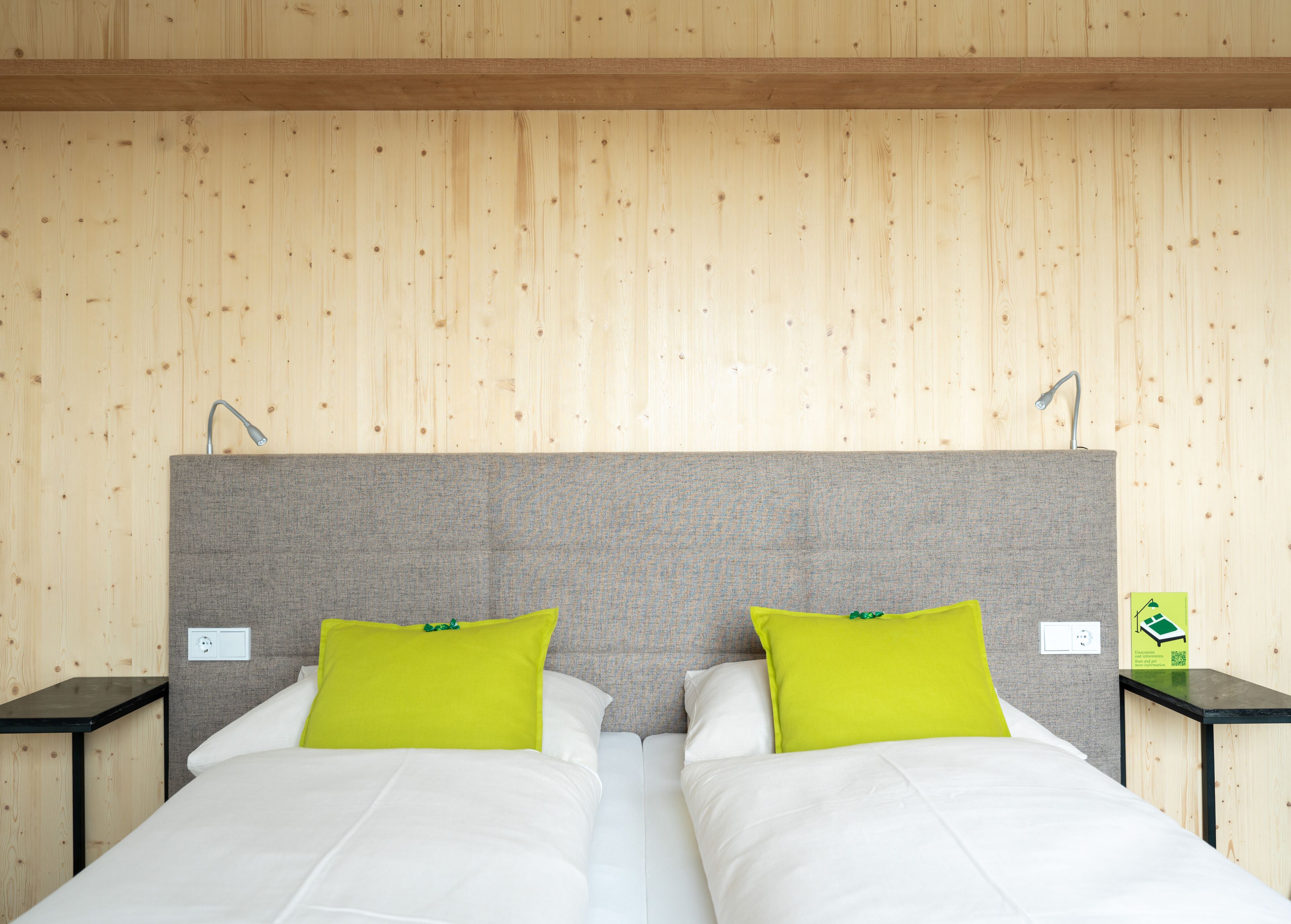 A double bed with white bedding and two bright green pillows, set against a wooden wall with a grey headboard.