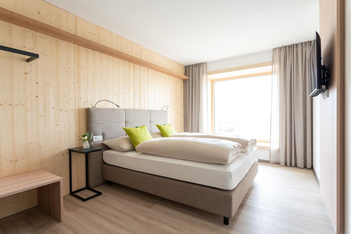 Une chambre moderne avec un mur en bois, un lit avec une literie blanche et des coussins verts, une table de chevet avec un vase et un téléviseur fixé au mur.
