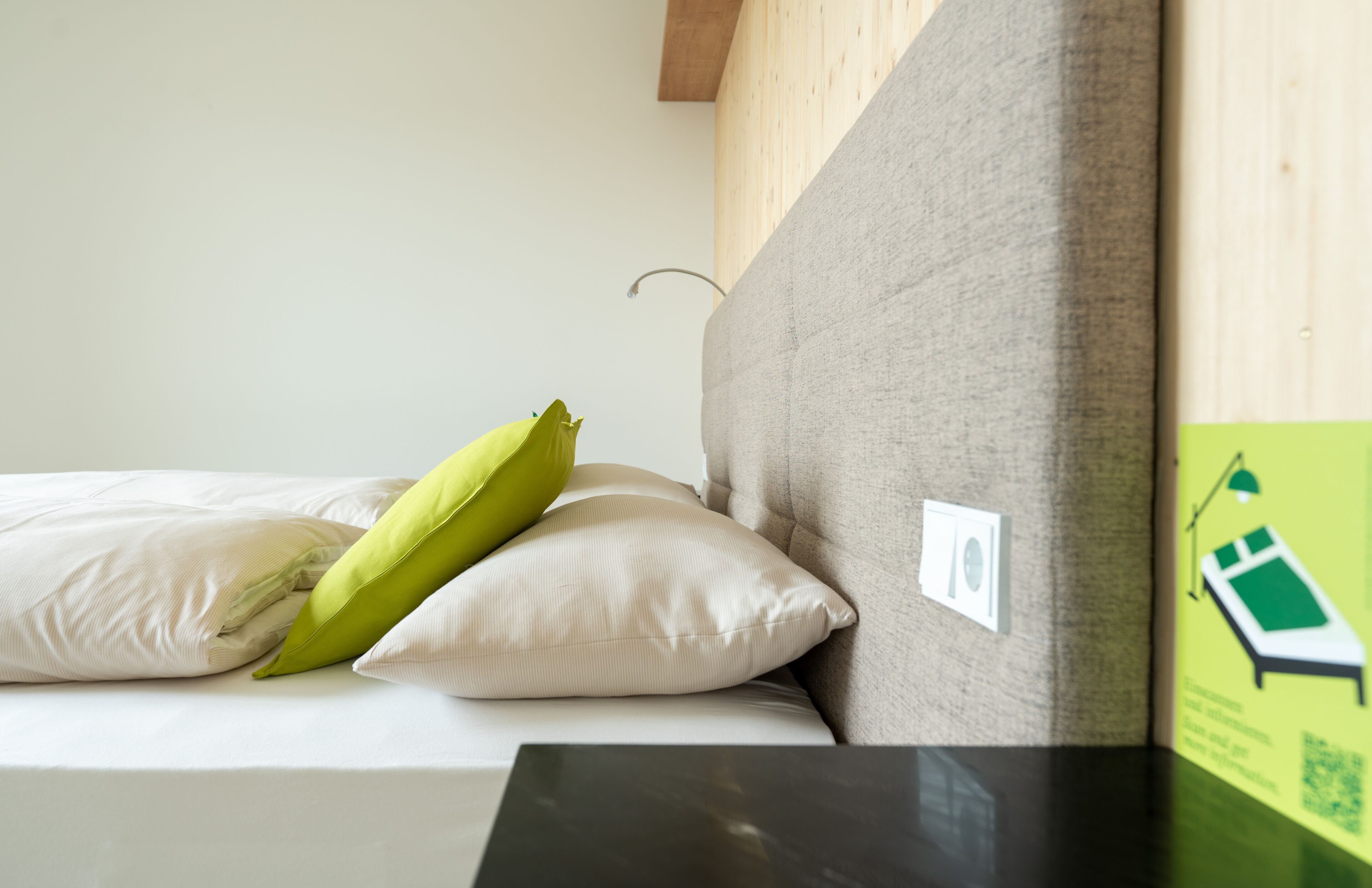 Uma cama com colchão bege, duas almofadas (uma verde), cabeceira de madeira e um interruptor branco pequeno na parede.