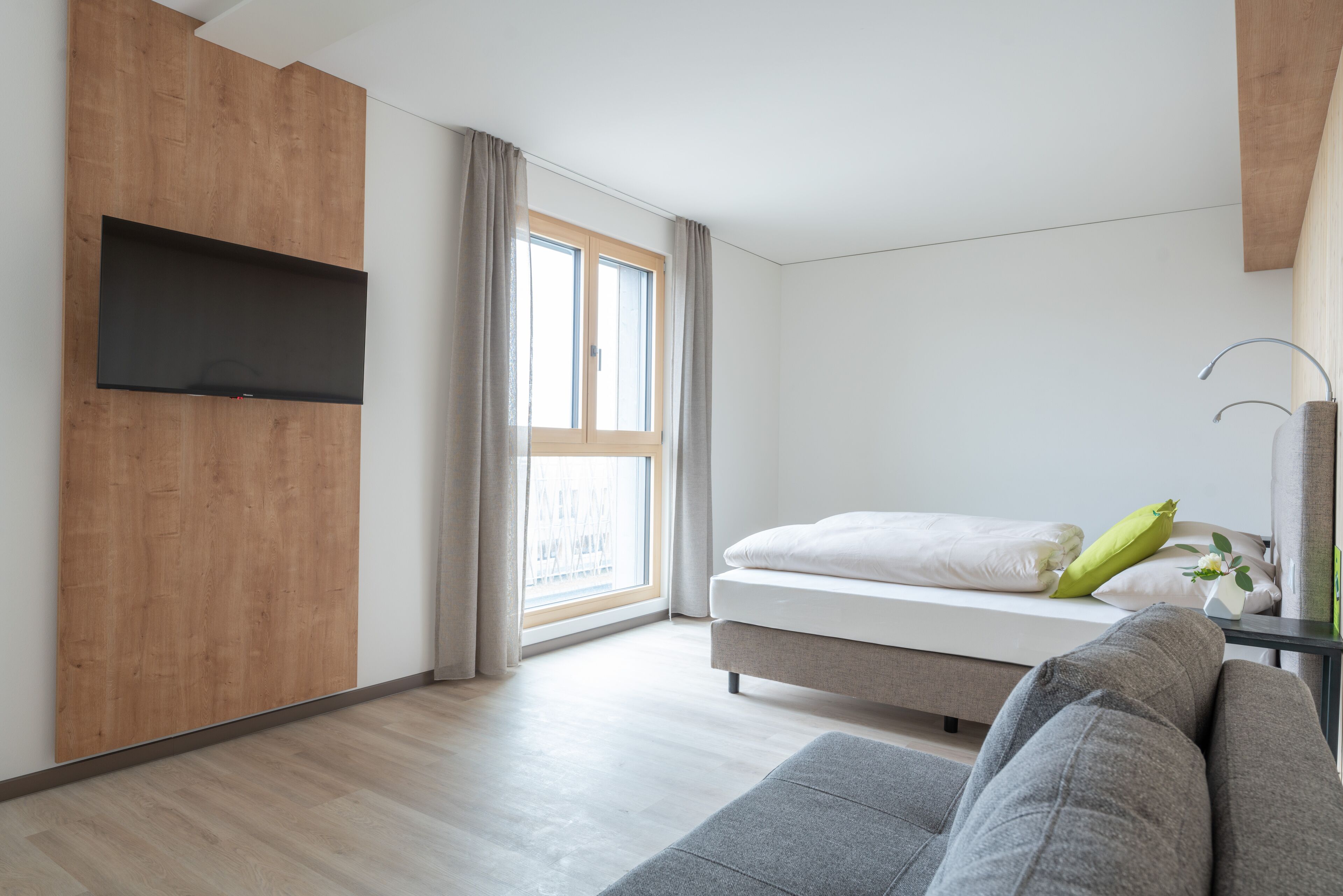 Un dormitorio moderno con una cama, un sofá gris, un televisor montado en un panel de madera y una ventana con cortinas.