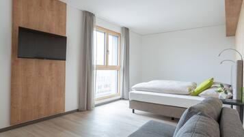 A modern bedroom with a bed, a grey sofa, a TV mounted on a wooden panel, and a window with curtains.