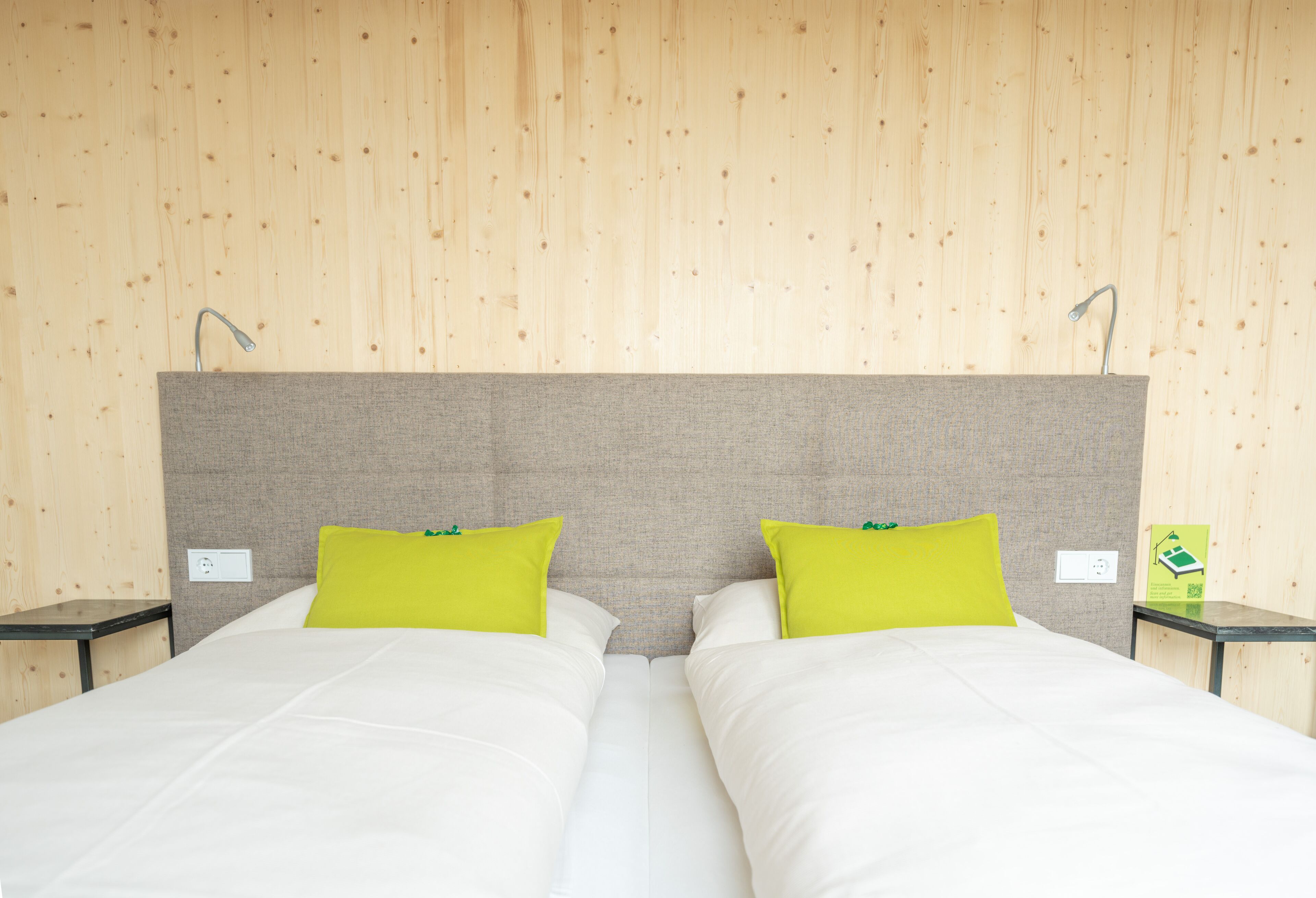 A double bed with white bedding and two bright green pillows, a grey headboard, and two bedside tables with lamps.