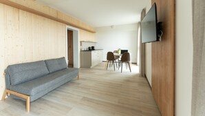 Una sala de estar moderna con un sofá gris, paneles de pared de madera, una zona de comedor con sillas y una mesa, y una cocina al fondo.