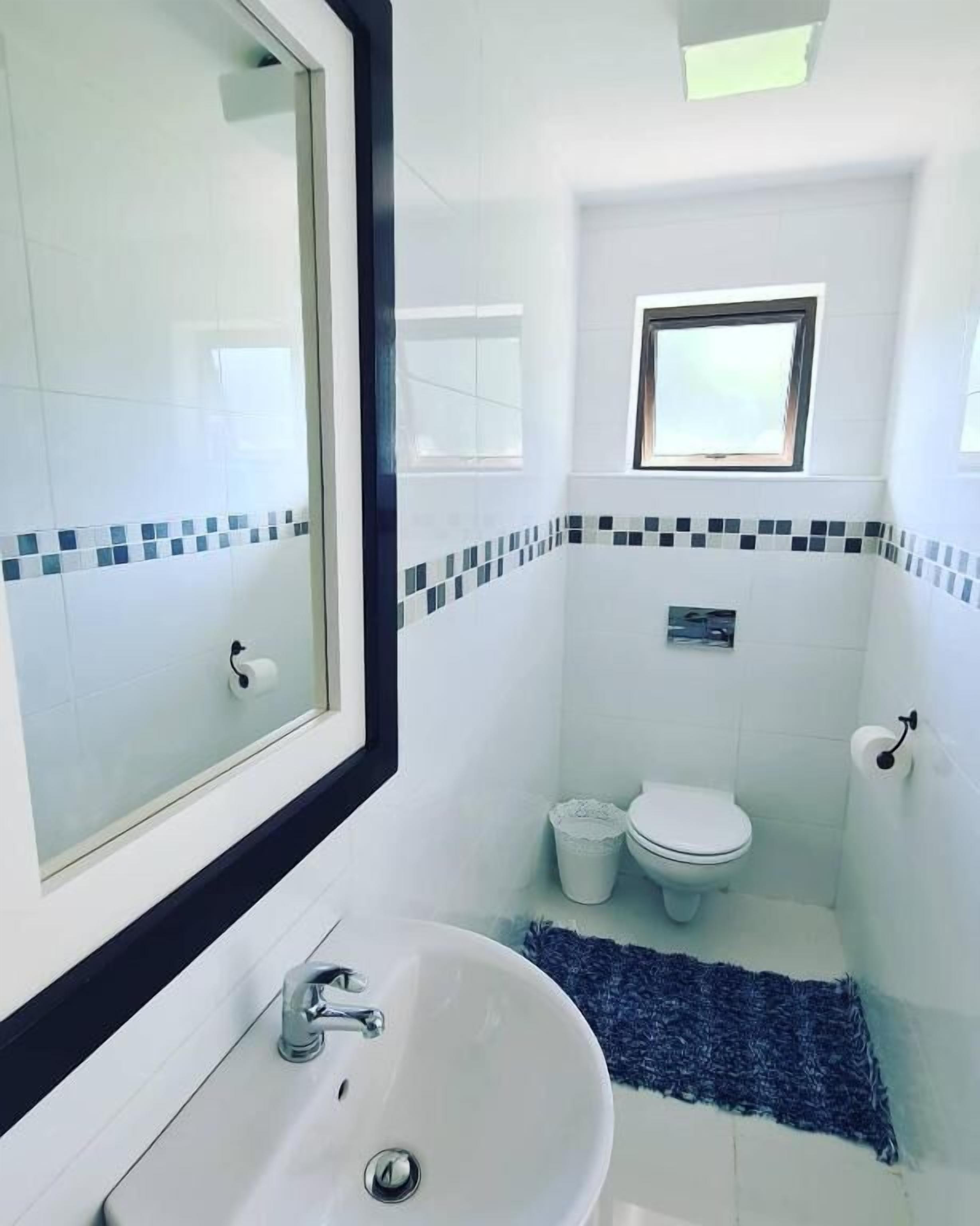 A small, clean bathroom with a white sink, a mirror, a toilet, and a window.