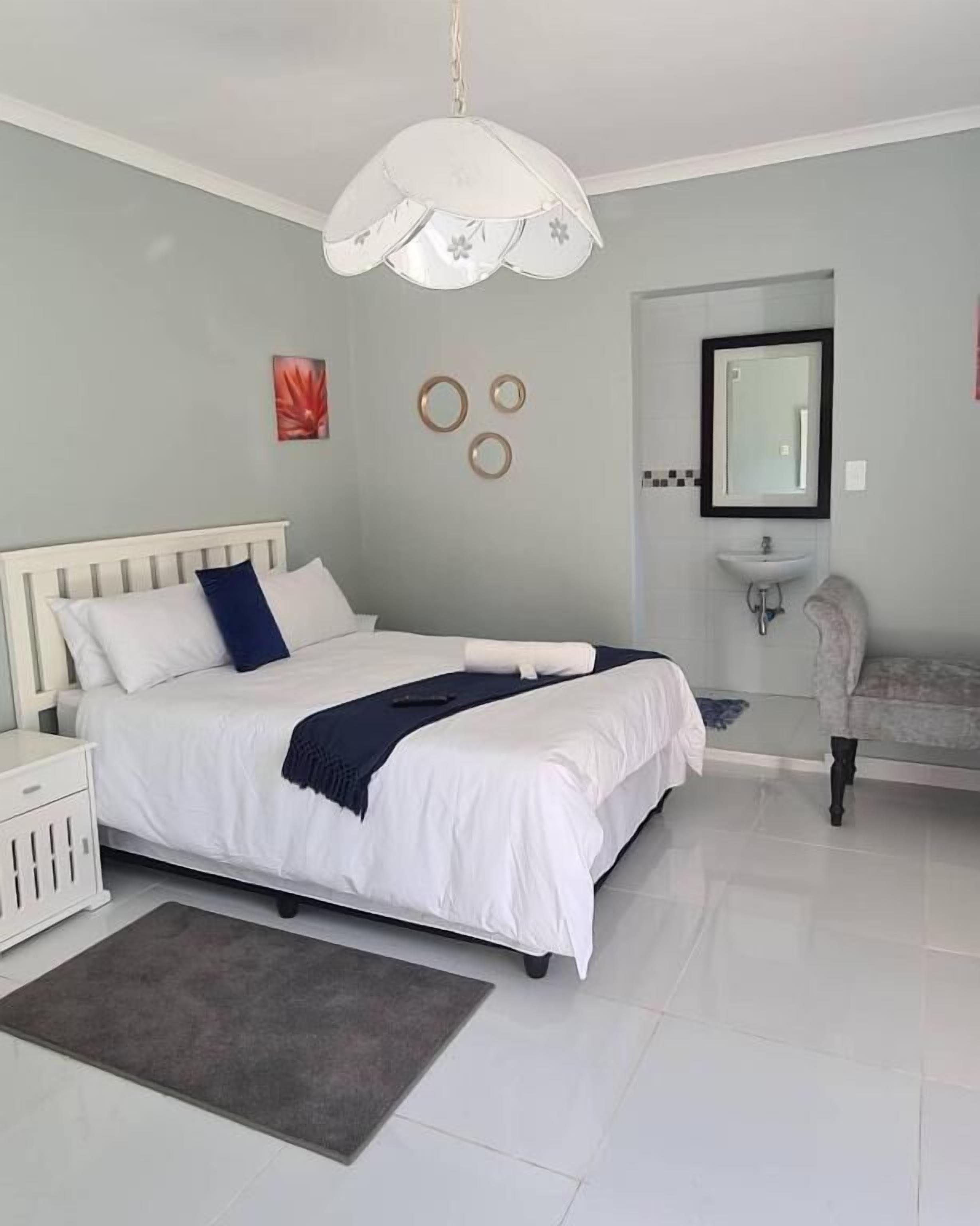Une chambre moderne avec un lit blanc, une table de chevet, un tapis gris et une salle de bain équipée d’un lavabo et d’un miroir.