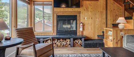 A cozy cabin interior with a fireplace, wooden walls, a TV mounted above it, and a view of the outdoors through large windows.
