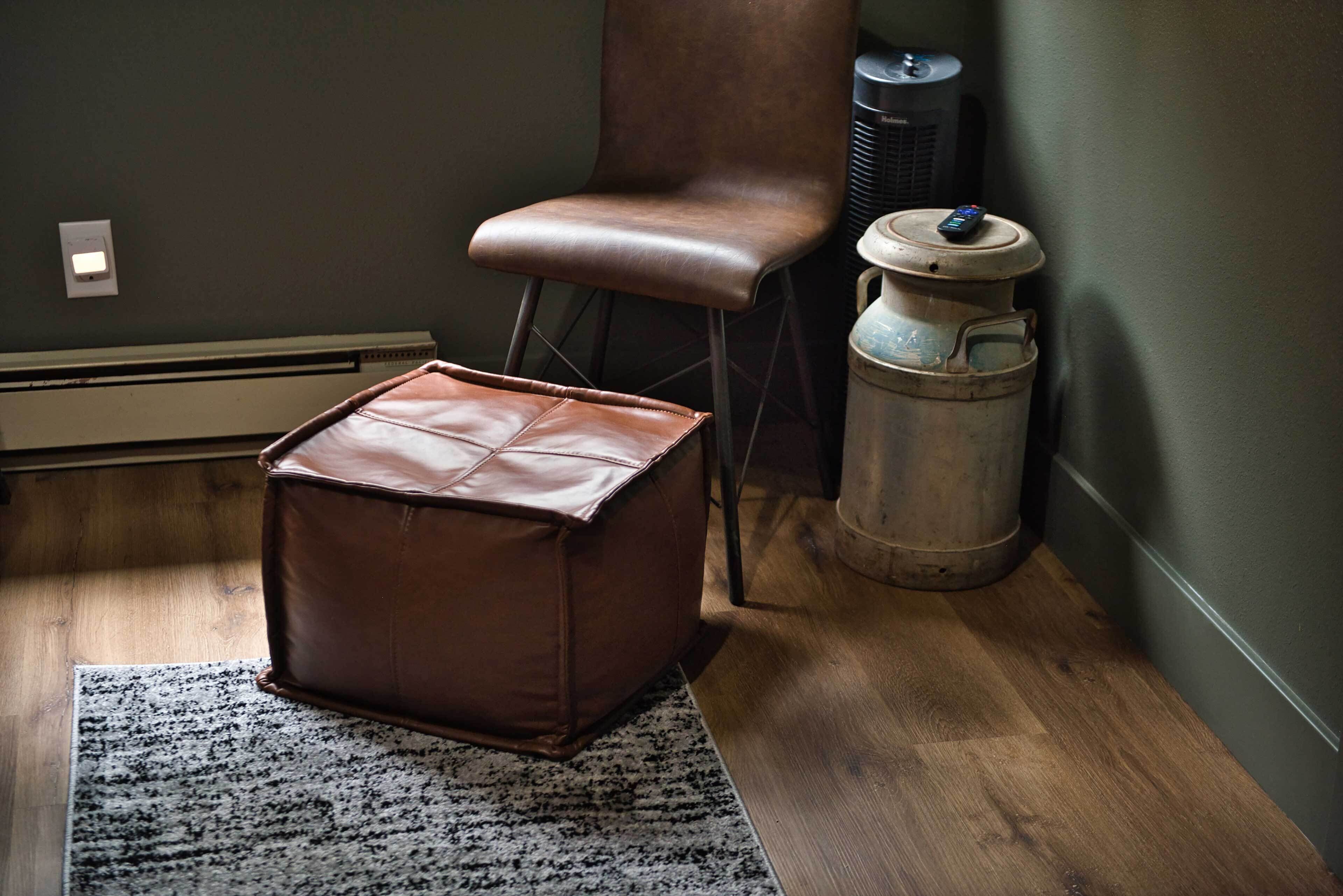 A room with a brown leather ottoman, a chair, a radiator, and a trash can.