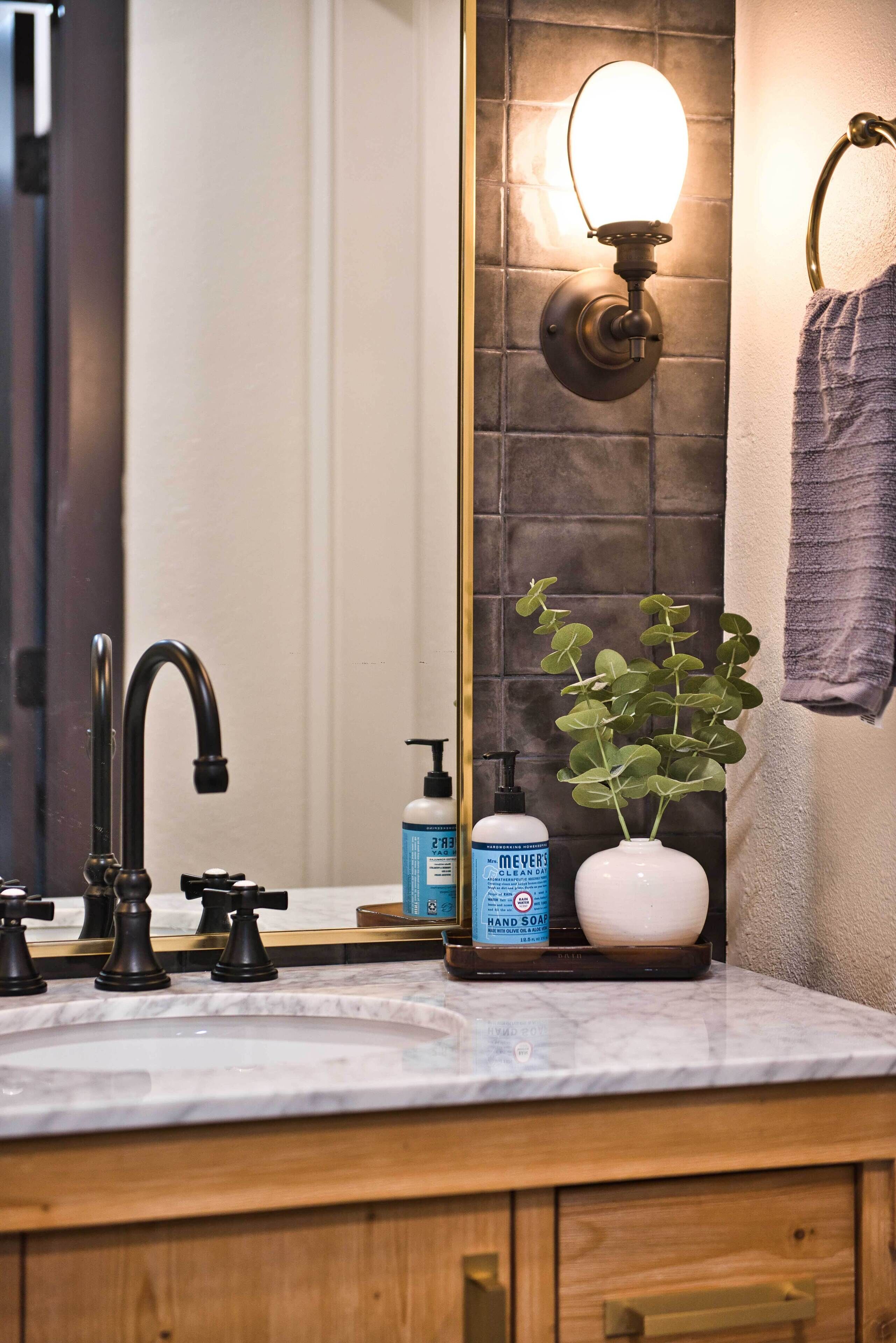 A bathroom with a marble sink, black faucets, a mirror with a wall-mounted light, a potted plant, and a towel rack.