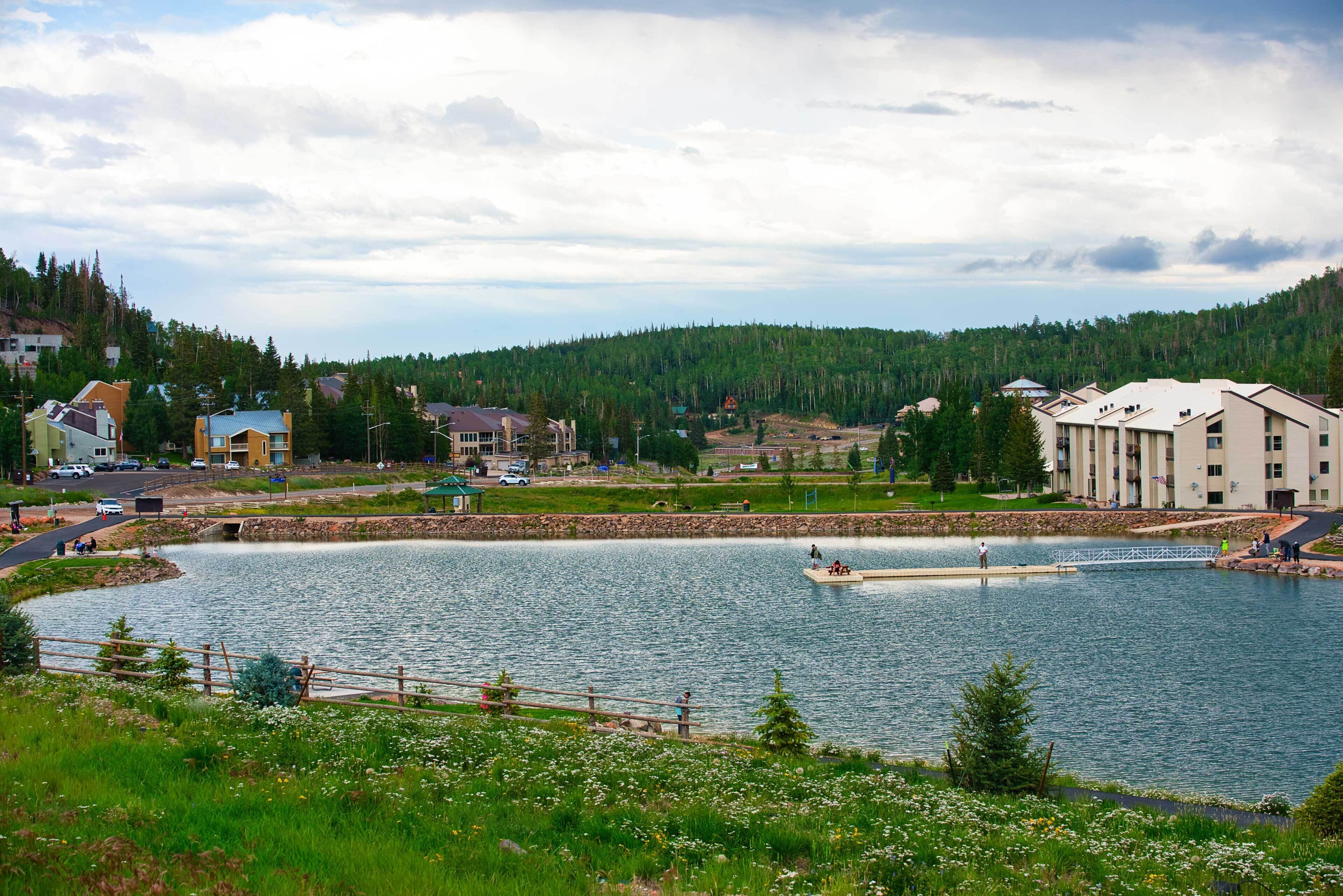 A lakeside view with a dock, buildings, and a forested hill.