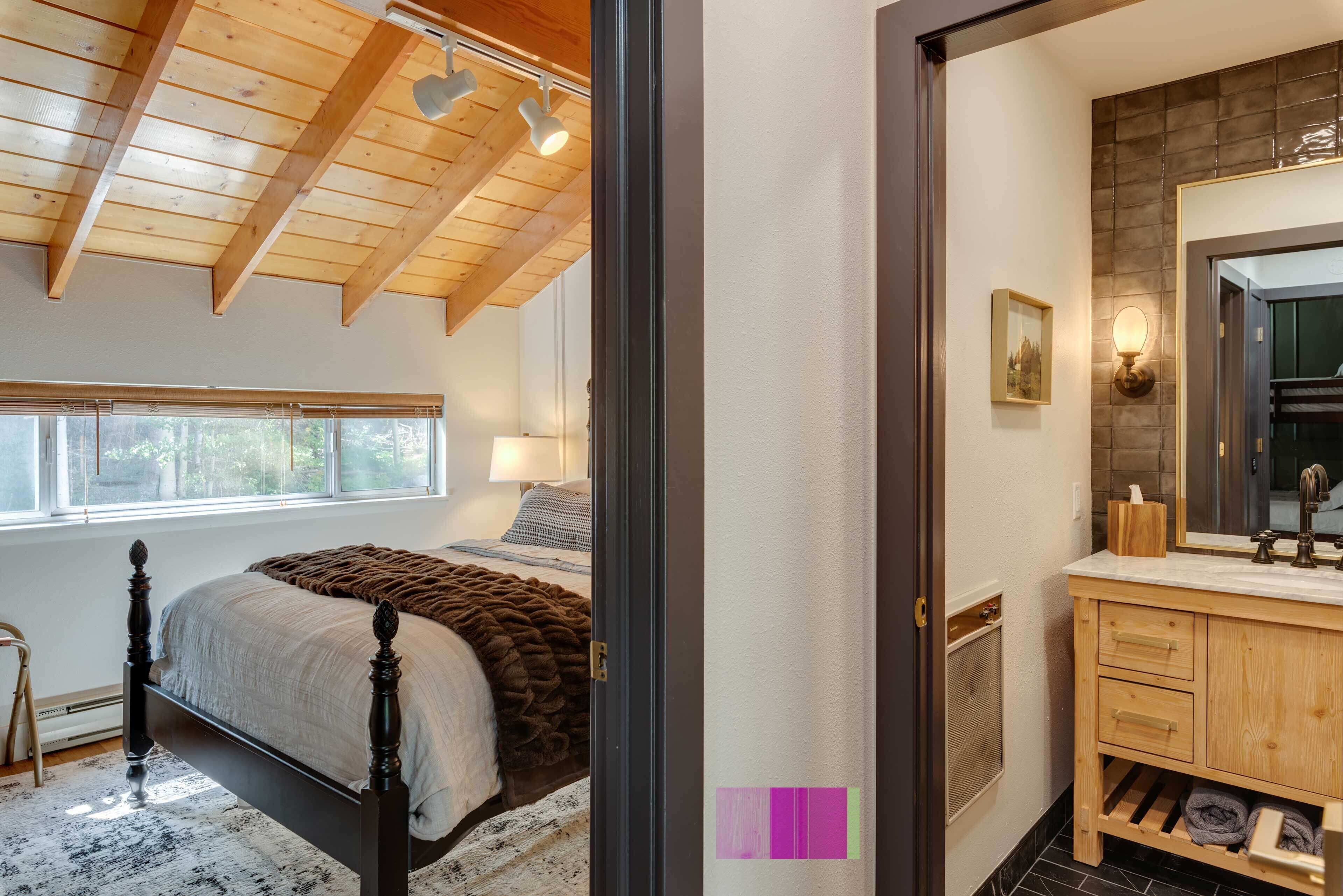 Un dormitorio con techo de madera, una cama grande, un ventanal y un baño con tocador y espejo de madera.