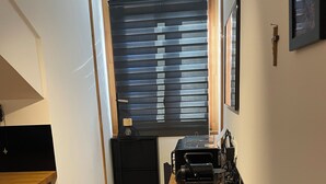 A compact office space with a wooden desk, a black filing cabinet, and a large window with blinds. - Studio MODERNE (Barcelonnette)