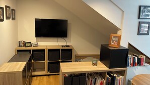 A modern living room with a flat-screen TV mounted on the wall, wooden shelving with books, a small table with a candle, and a staircase with a view to the upper floor. - Studio MODERNE (Barcelonnette)