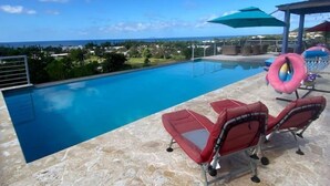 A swimming pool with a view of the ocean, surrounded by lounge chairs and umbrellas. - Ocean View Villa with Infinity Pool, Hot Tub & Fire Pit; 5 Min from Beach (Rincón)