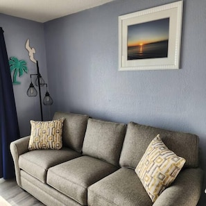 A living room with a grey sofa, patterned cushions, a framed picture of a sunset, and decorative wall art.